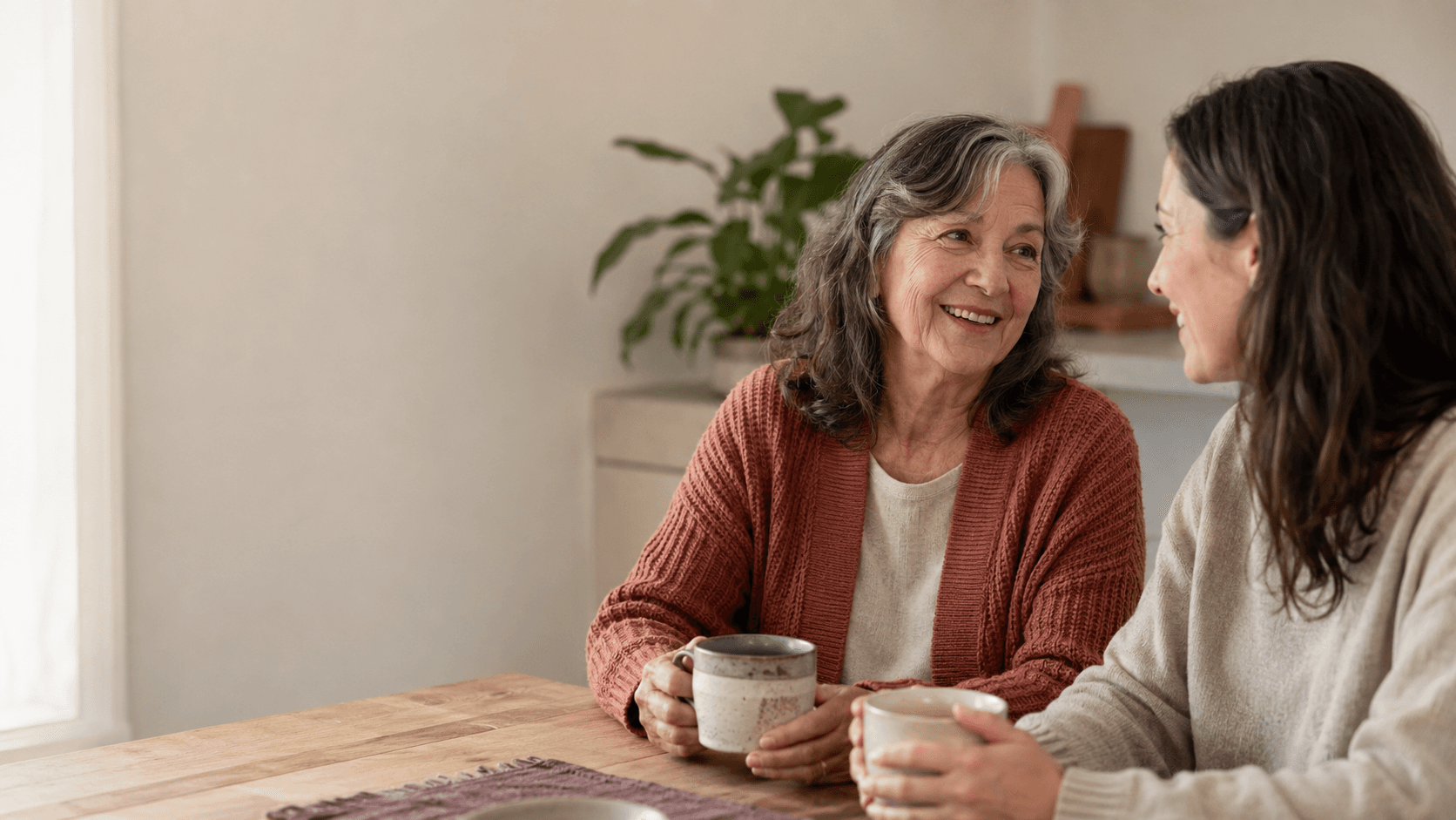 An adult daughter and her mother sitting together at a sunlit kitchen table, sharing a warm conversation.